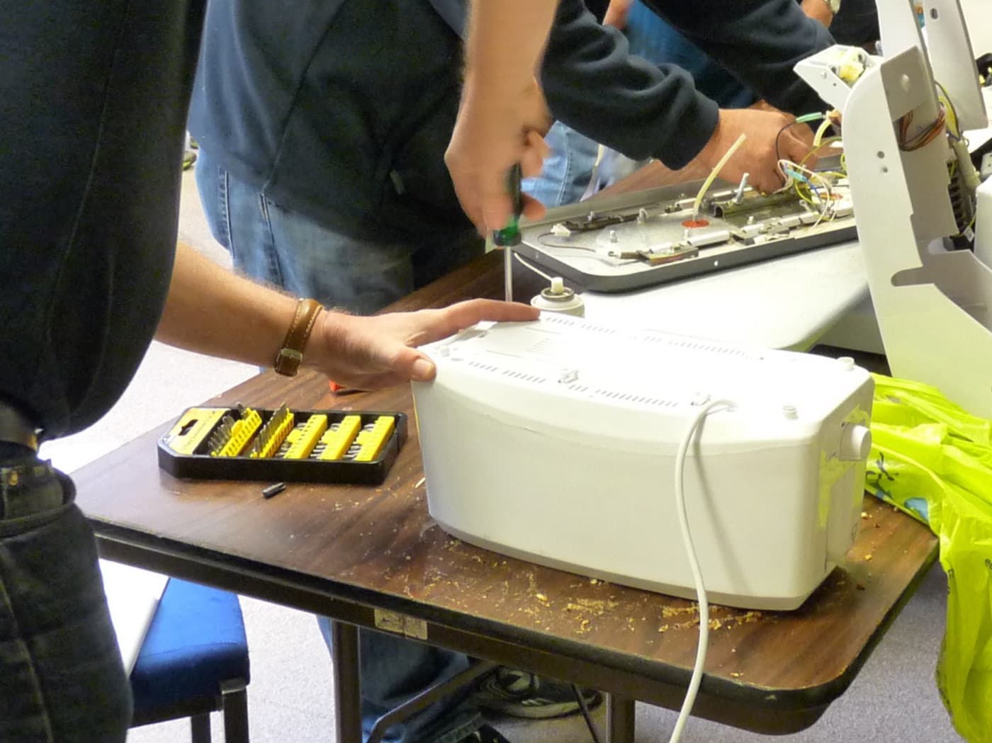 Deux paires de mains autour d’une machine à coudre ouverte sur une table d’atelier, un tournevis dans le capot et une trousse d’embouts posée à côté.